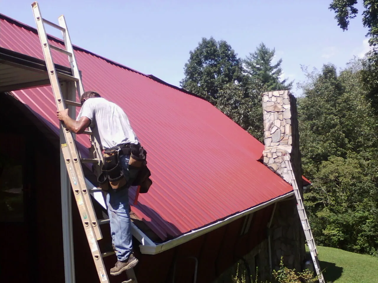 Roofer on a ladder beside a red metal roof and stone chimney — Soffit & Fascia Repair by Roofing Brothers in Roaring Gap